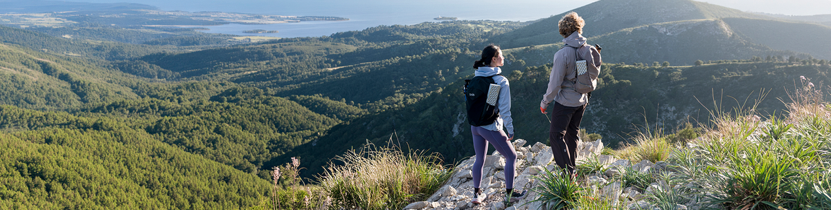 Two hikers with backpacks looking out over forested hills and the sea